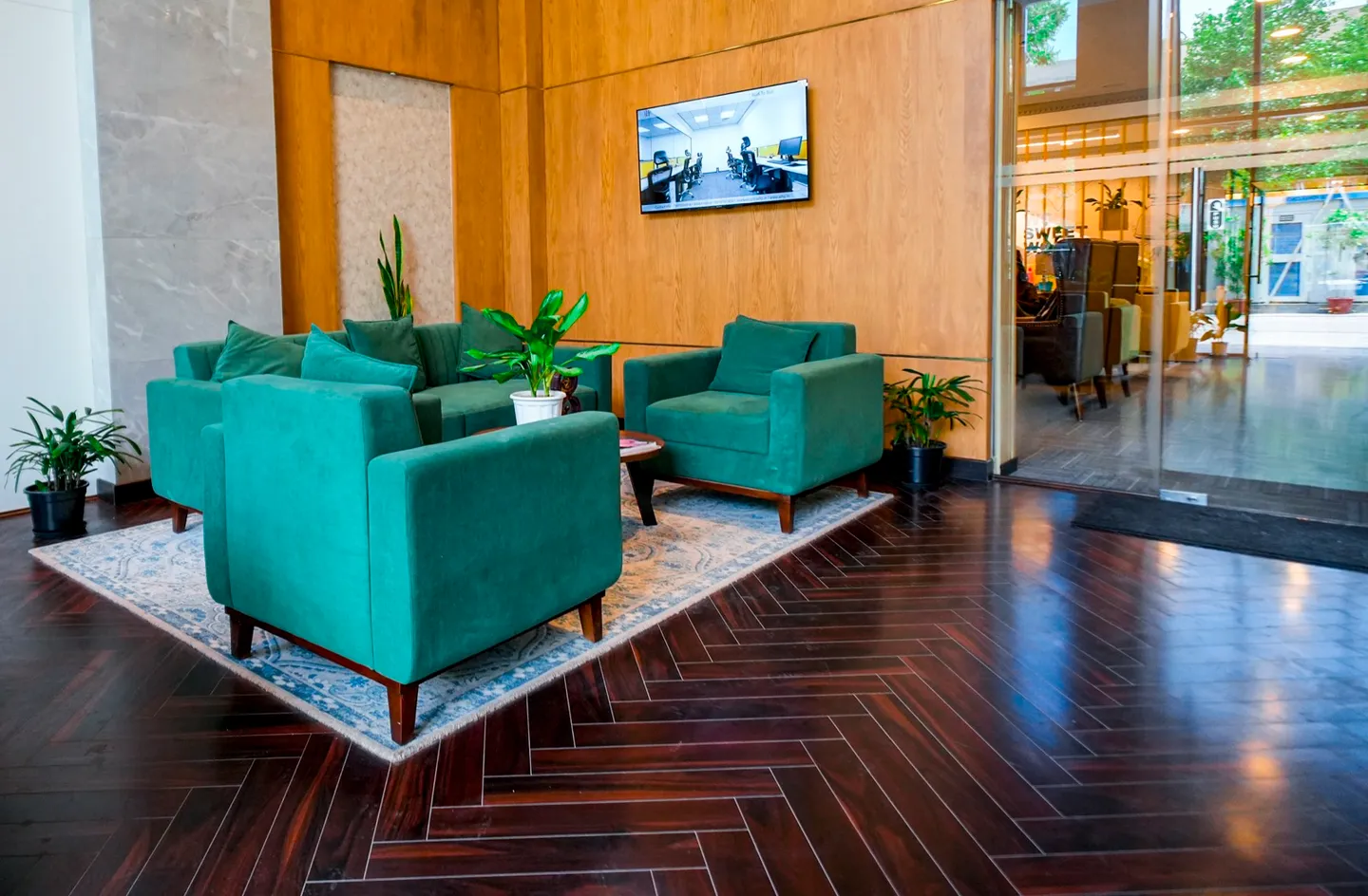 Teal velvet armchairs on a blue rug over dark herringbone wood floor by a glass entrance wall — AIHP managed office, Gurgaon