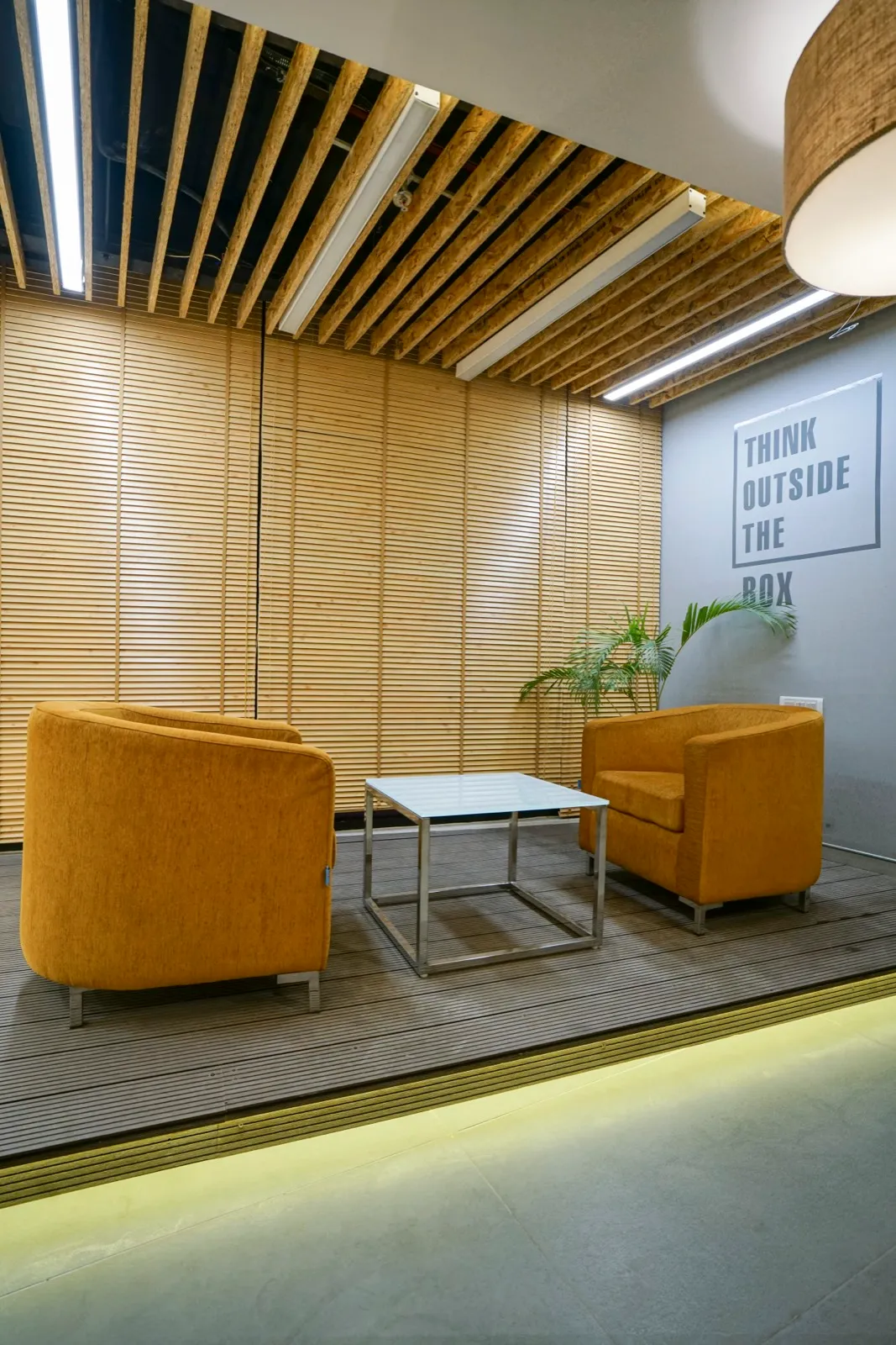 Mustard armchairs and chrome cube table on raised wood deck with cove lighting and OSB slat ceiling — AIHP managed office, Gurgaon