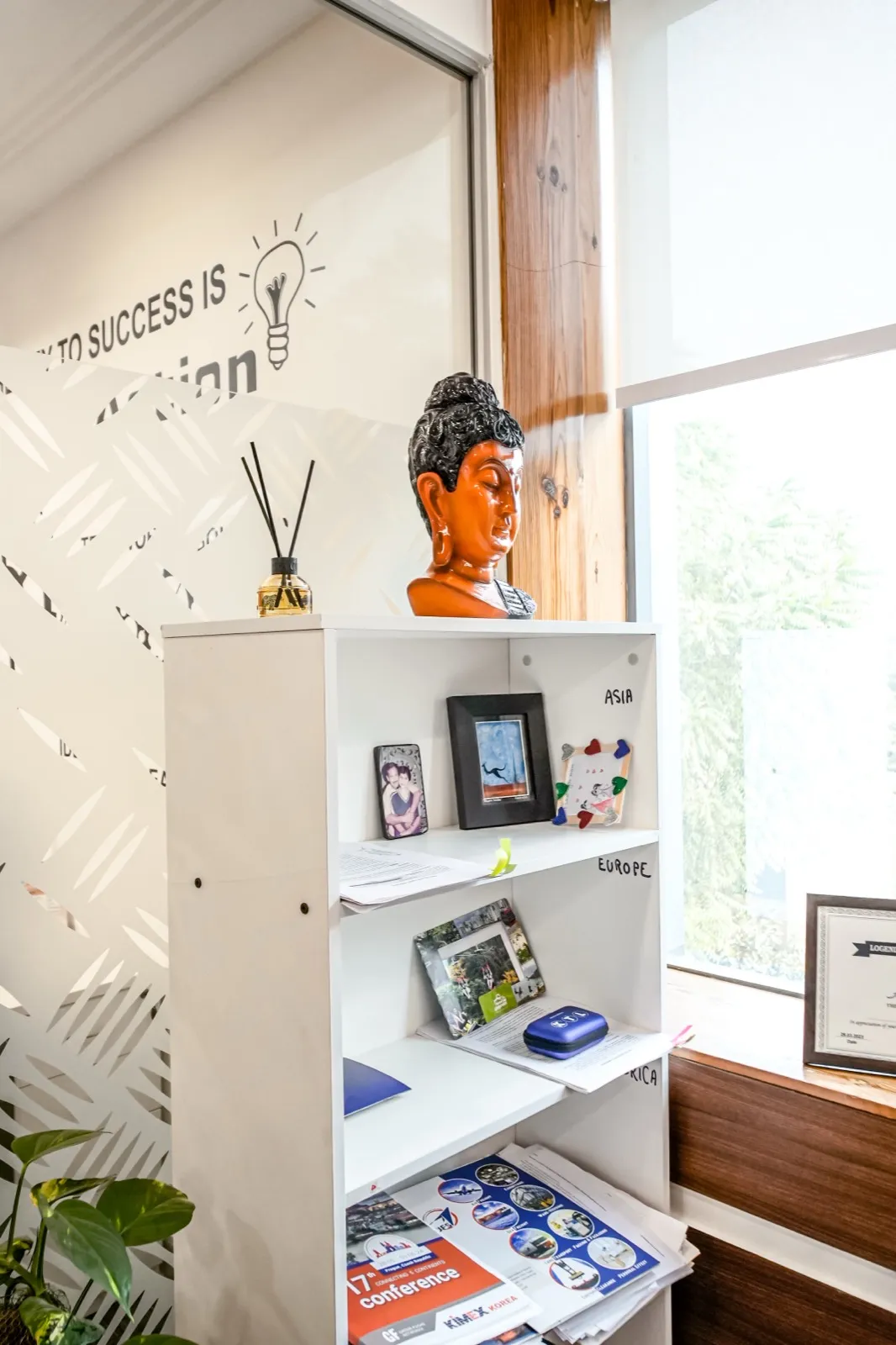 White bookshelf topped with orange Buddha bust and reed diffuser beside a sunlit timber-framed window — AIHP managed office, Gurgaon