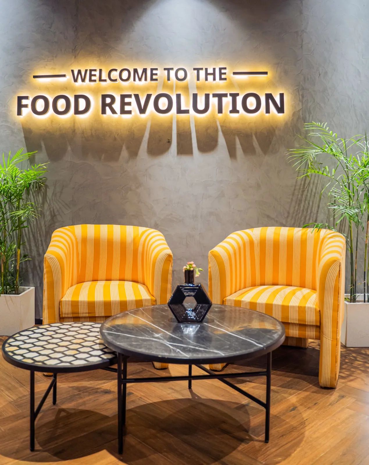 Yellow-striped tub chairs flanking marble and mosaic round tables under a backlit Food Revolution sign — AIHP managed office, Gurgaon