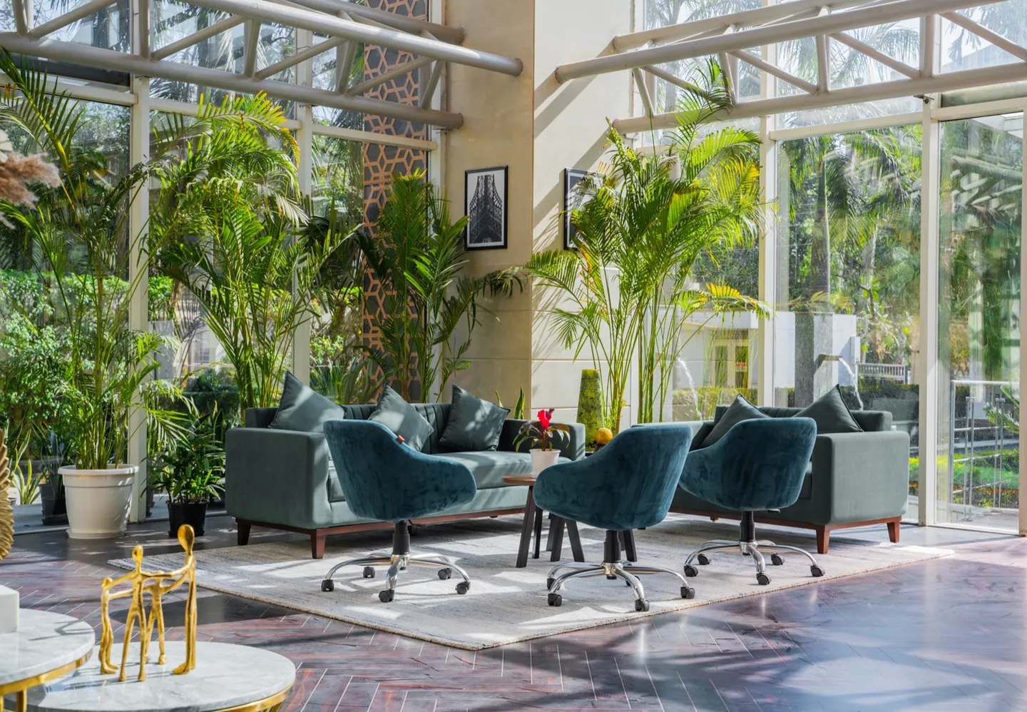 Teal velvet sofas and swivel chairs amid tall palms in a glass-walled atrium with marble floor — AIHP managed office, Gurgaon