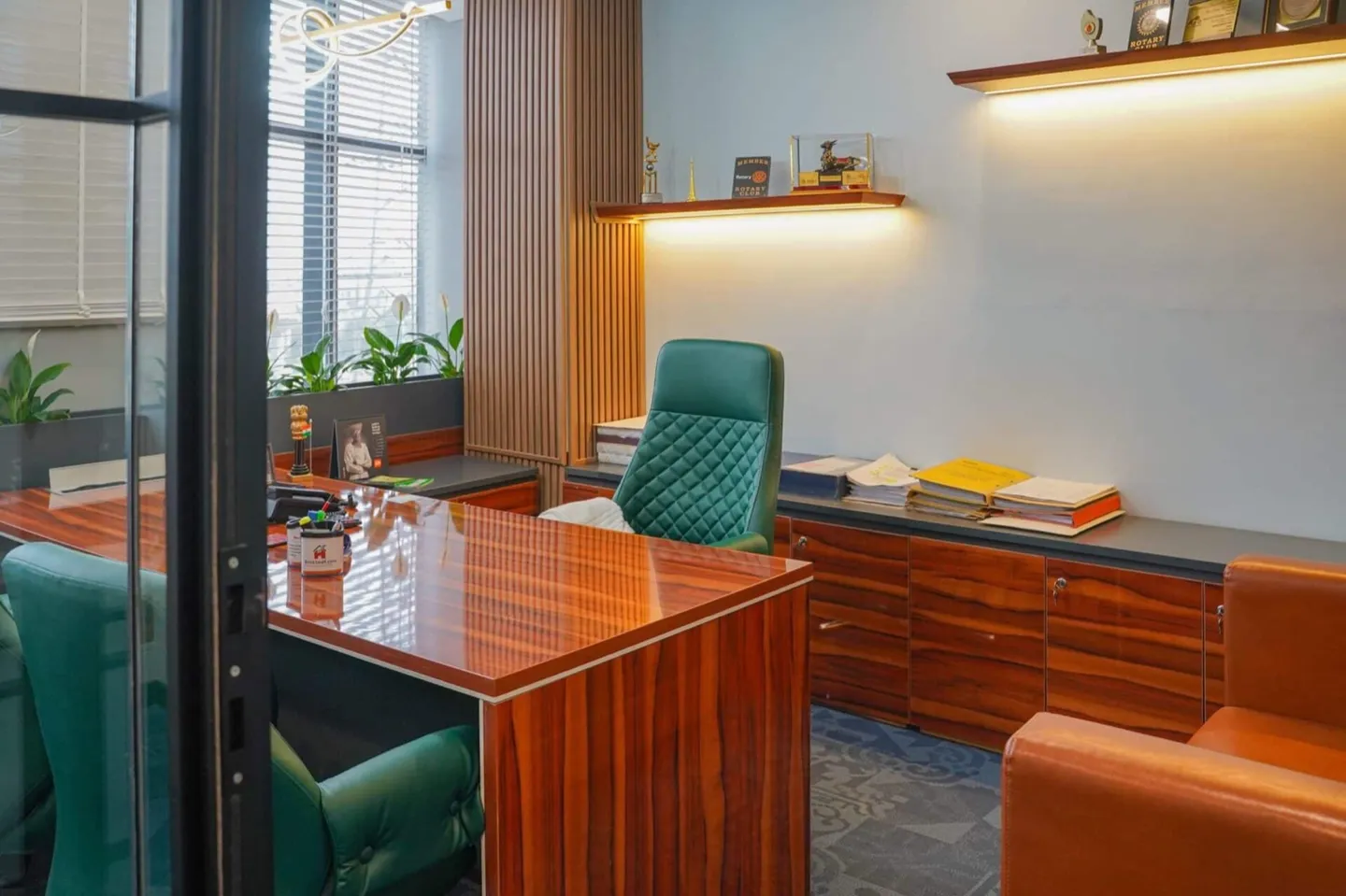 Glass-door cabin with glossy wood desk, green tufted leather chairs and a backlit trophy shelf — AIHP managed office, Gurgaon