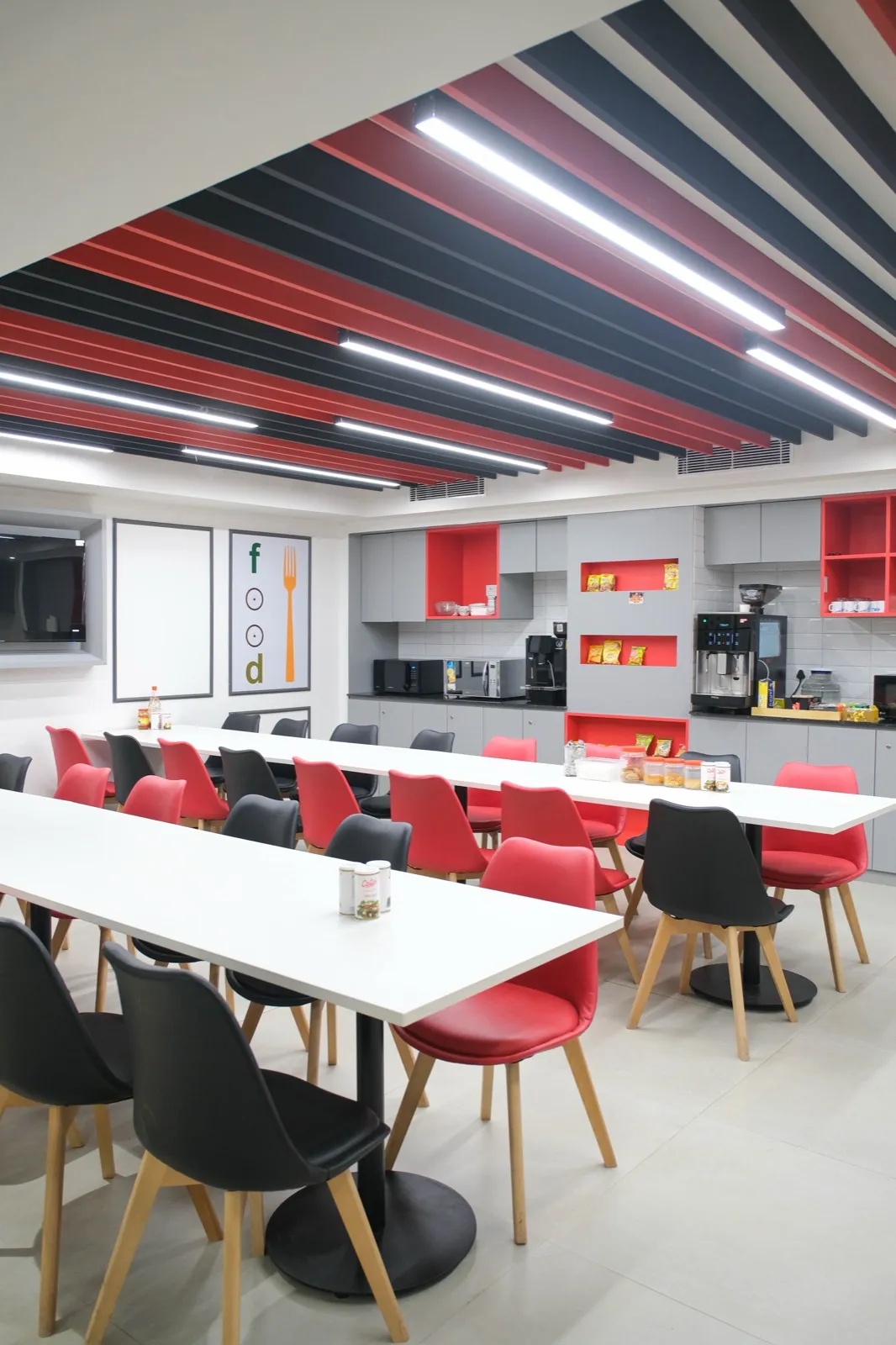 Staff cafeteria with long white tables, red and black chairs and a grey pantry counter with coffee machines — AIHP managed office, Gurgaon