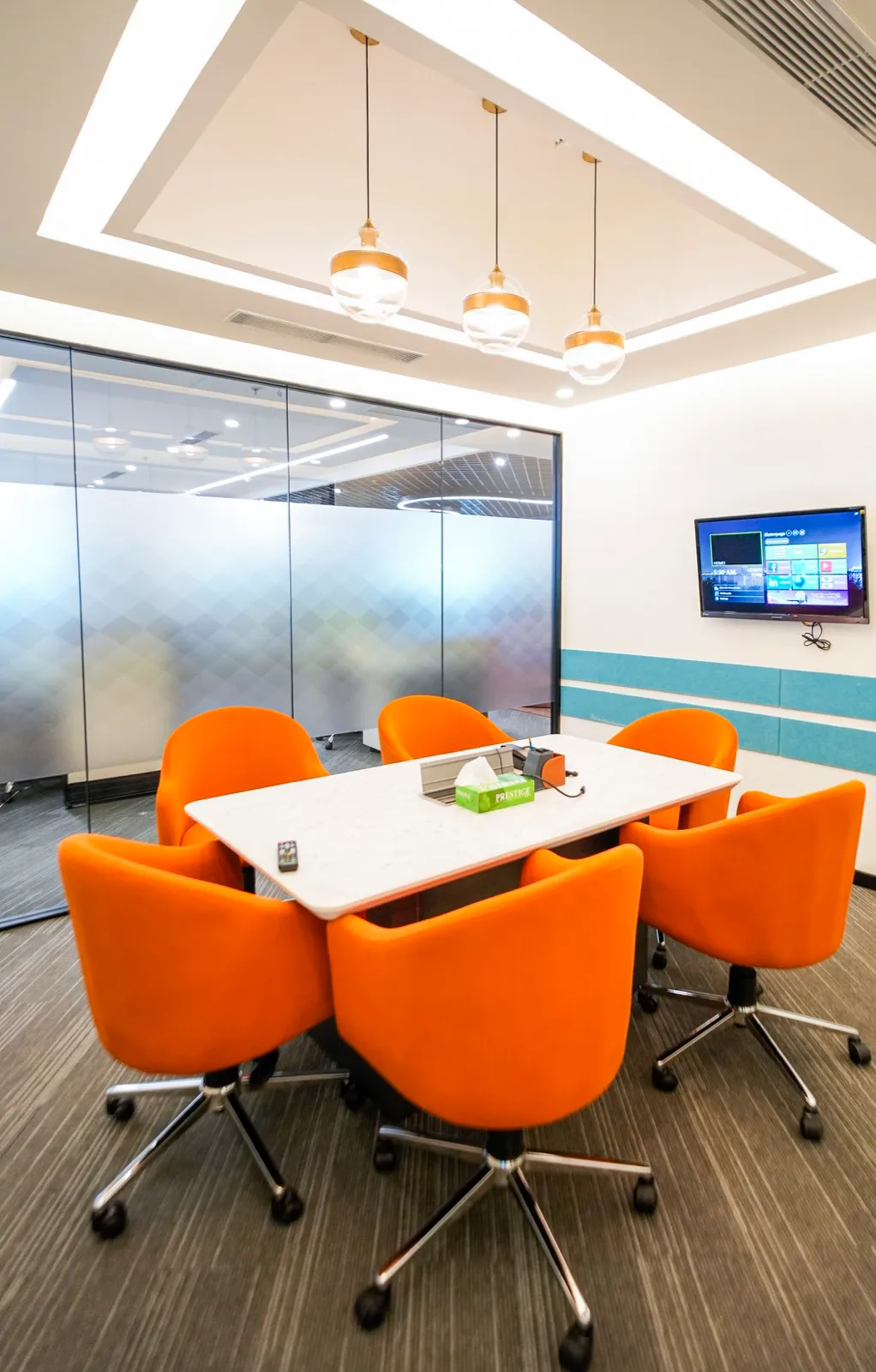 Six orange tub chairs around a white stone-top table by a frosted glass wall and screen — AIHP managed office, Gurgaon
