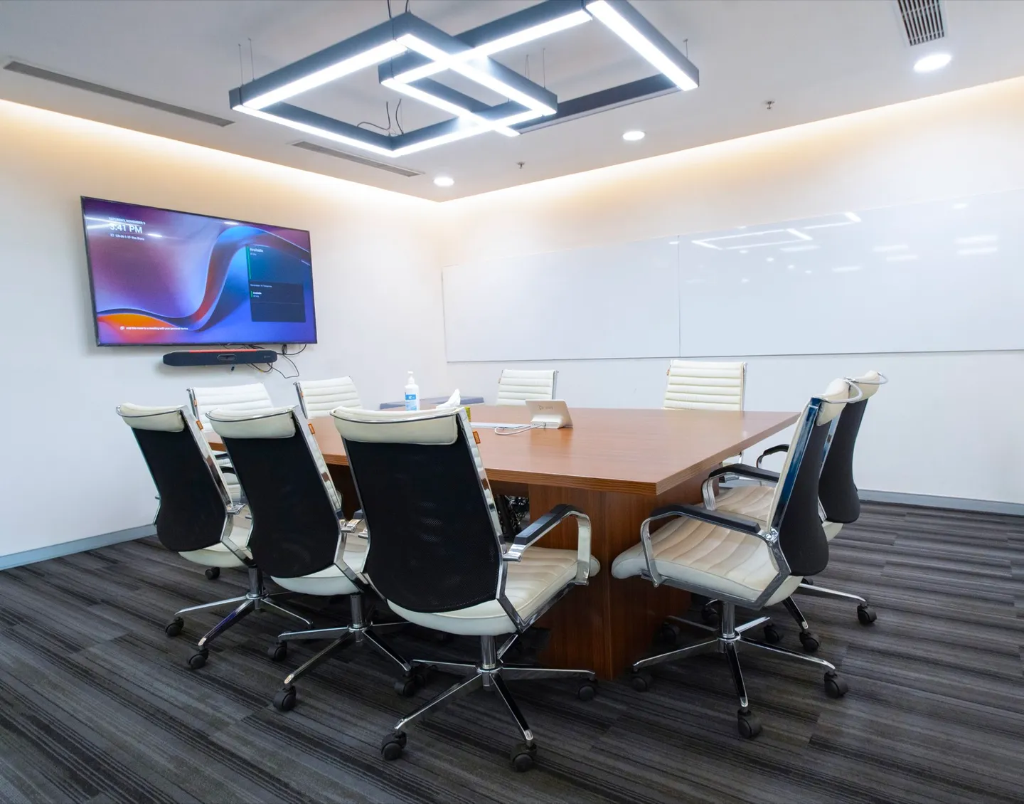 Eight cream-and-black chairs around a wood table facing a wall TV and whiteboard — AIHP managed office, Gurgaon