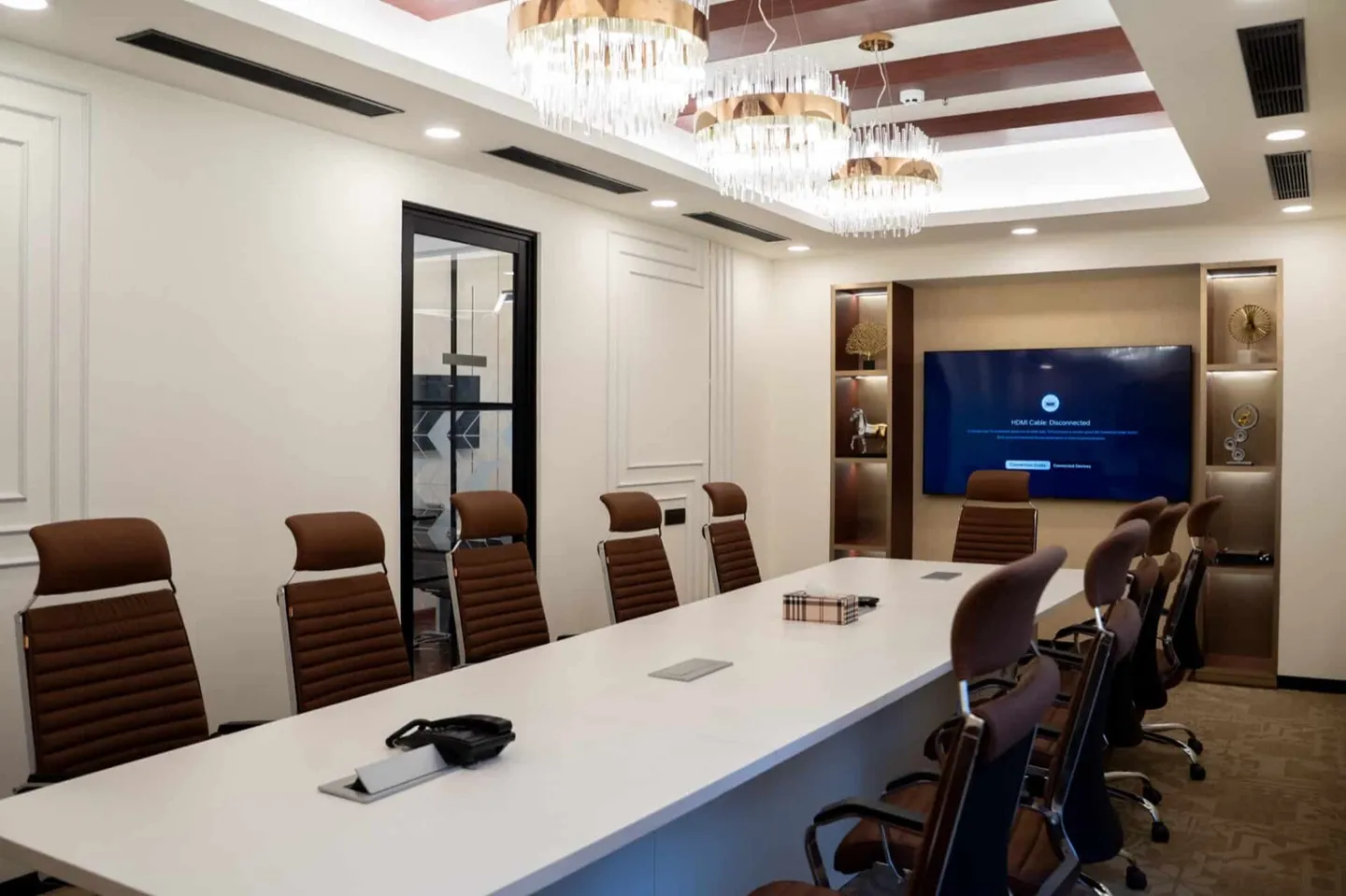 Twelve-seat white table with tan ribbed chairs, crystal chandeliers and a recessed display — AIHP managed office, Gurgaon