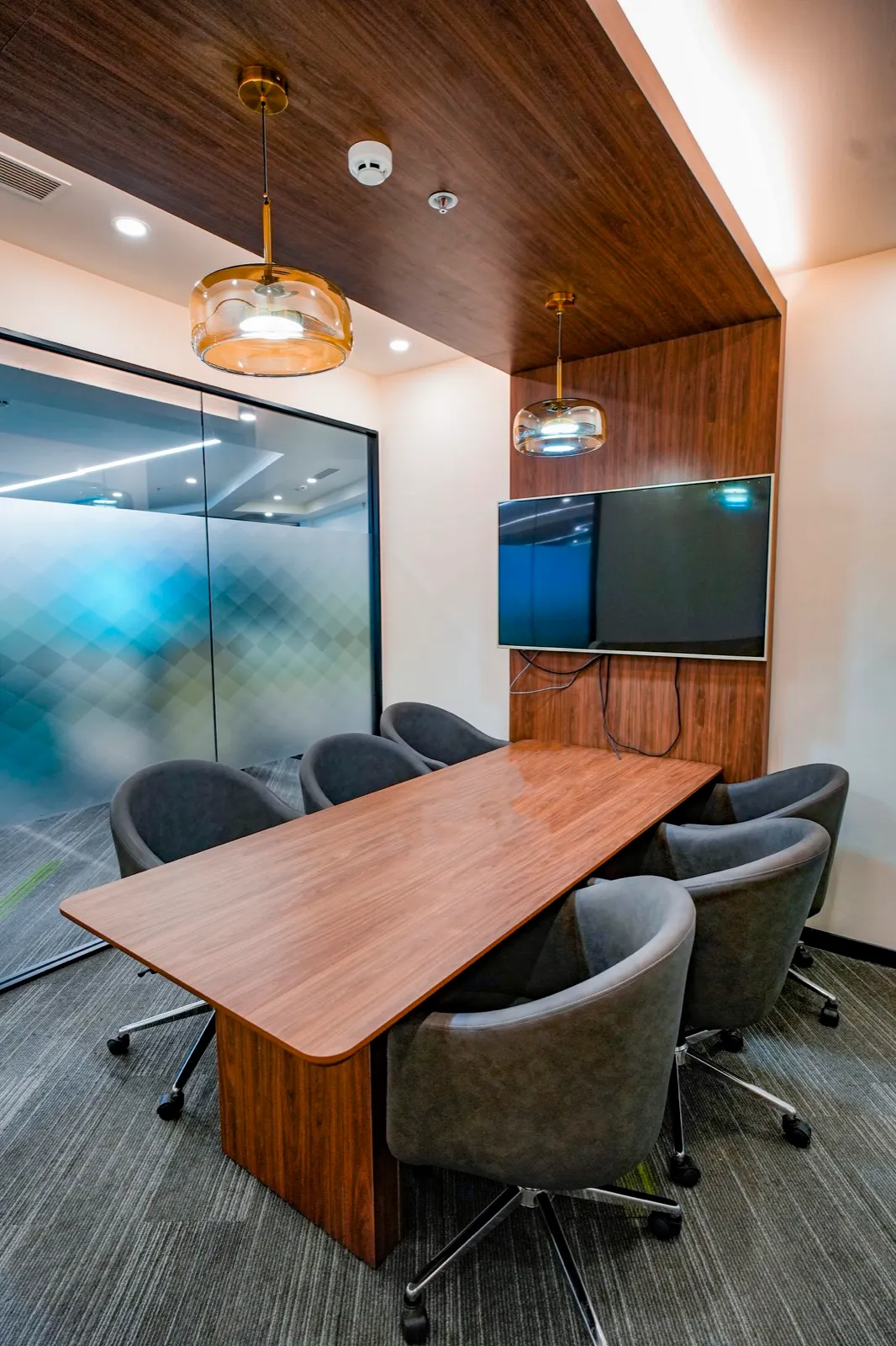 Six grey fabric chairs at a walnut table facing a wall TV on wood panelling — AIHP managed office, Gurgaon