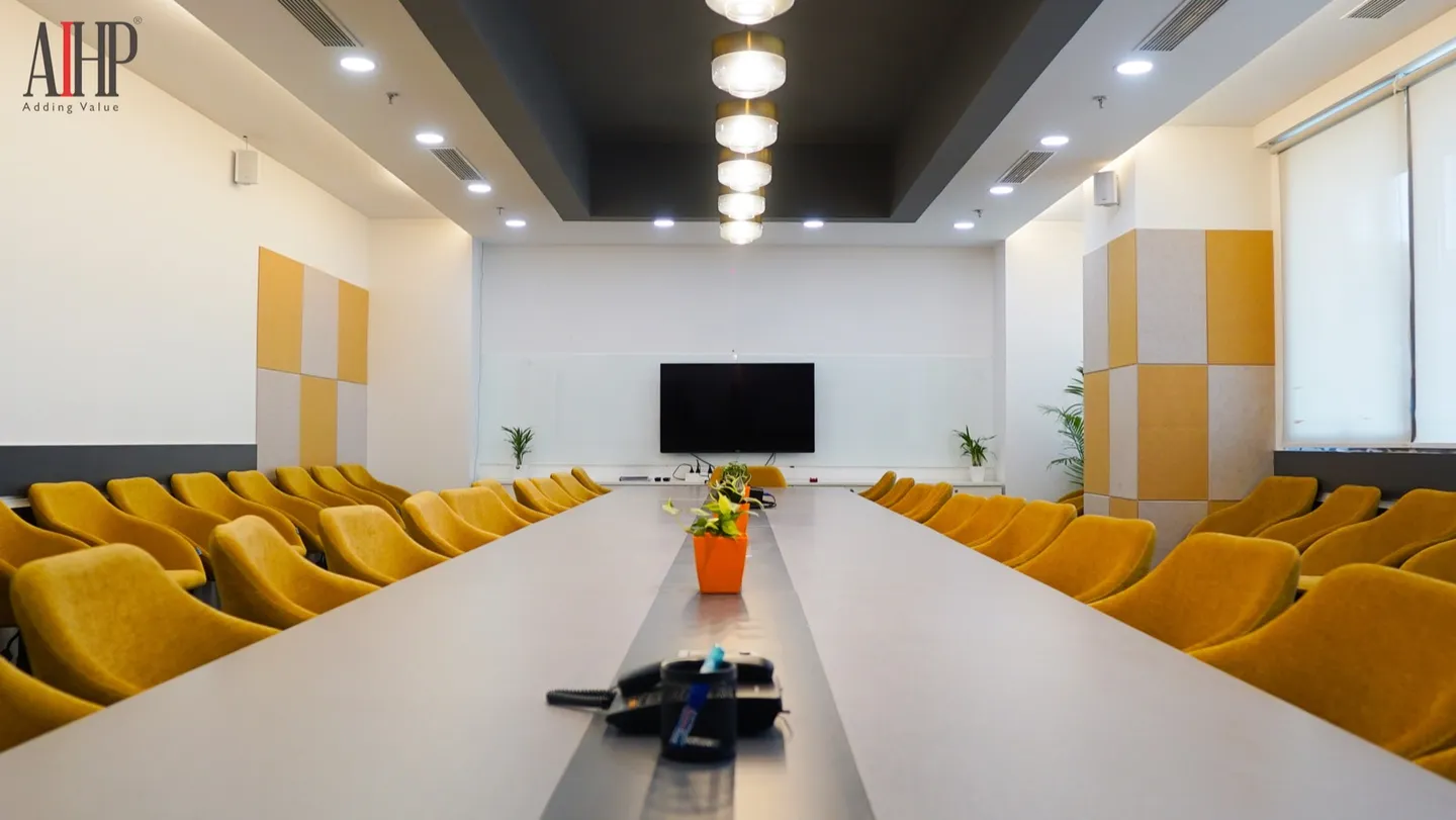 Long white conference table seating 30-plus in mustard armchairs facing a wall-mounted screen, pendant lights above — AIHP managed office, Gurgaon