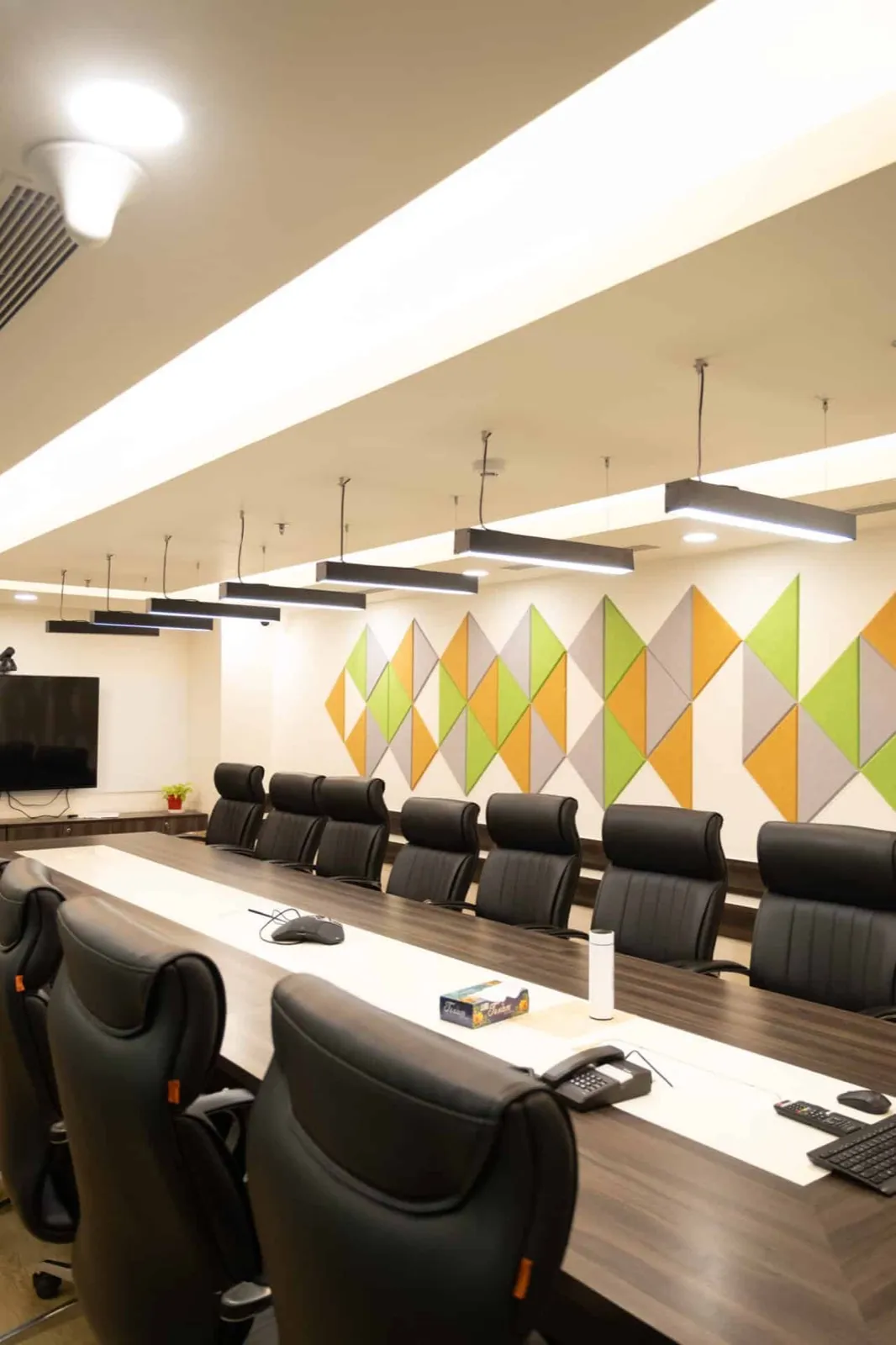 Boardroom with two-tone wood table, black executive chairs and orange-green triangular felt acoustic wall — AIHP managed office, Gurgaon