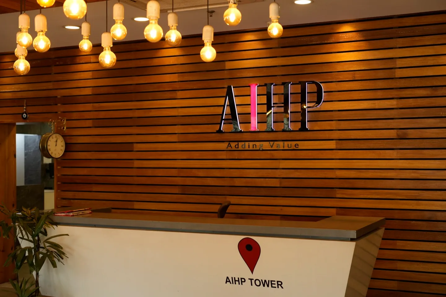 Wood-clad AIHP reception with the backlit AIHP 'Adding Value' logo, Edison-bulb pendant cluster and AIHP Tower welcome desk in Gurgaon