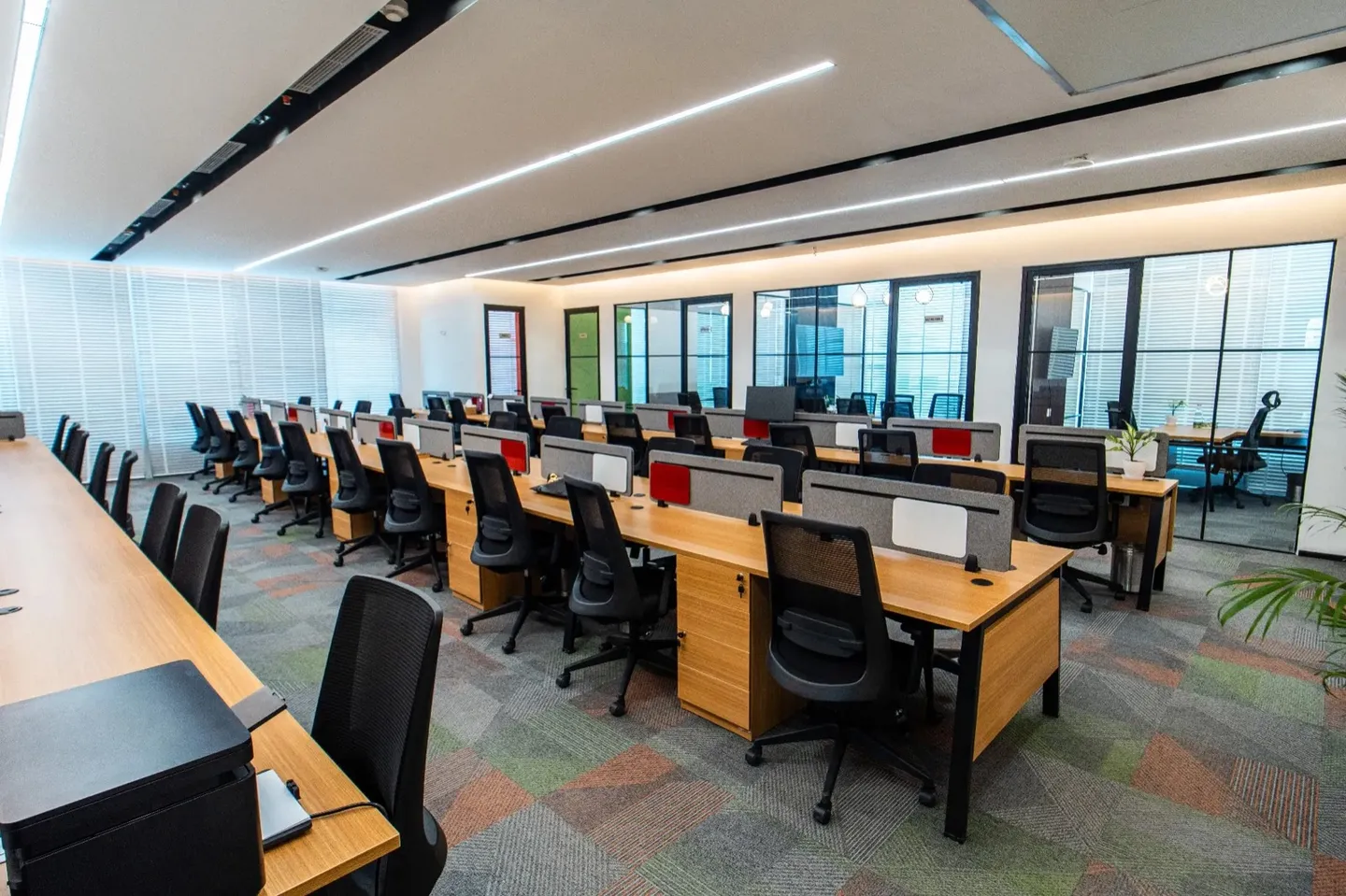 Open desk rows with black mesh chairs, red panels and glass cabins behind — AIHP managed office, Gurgaon