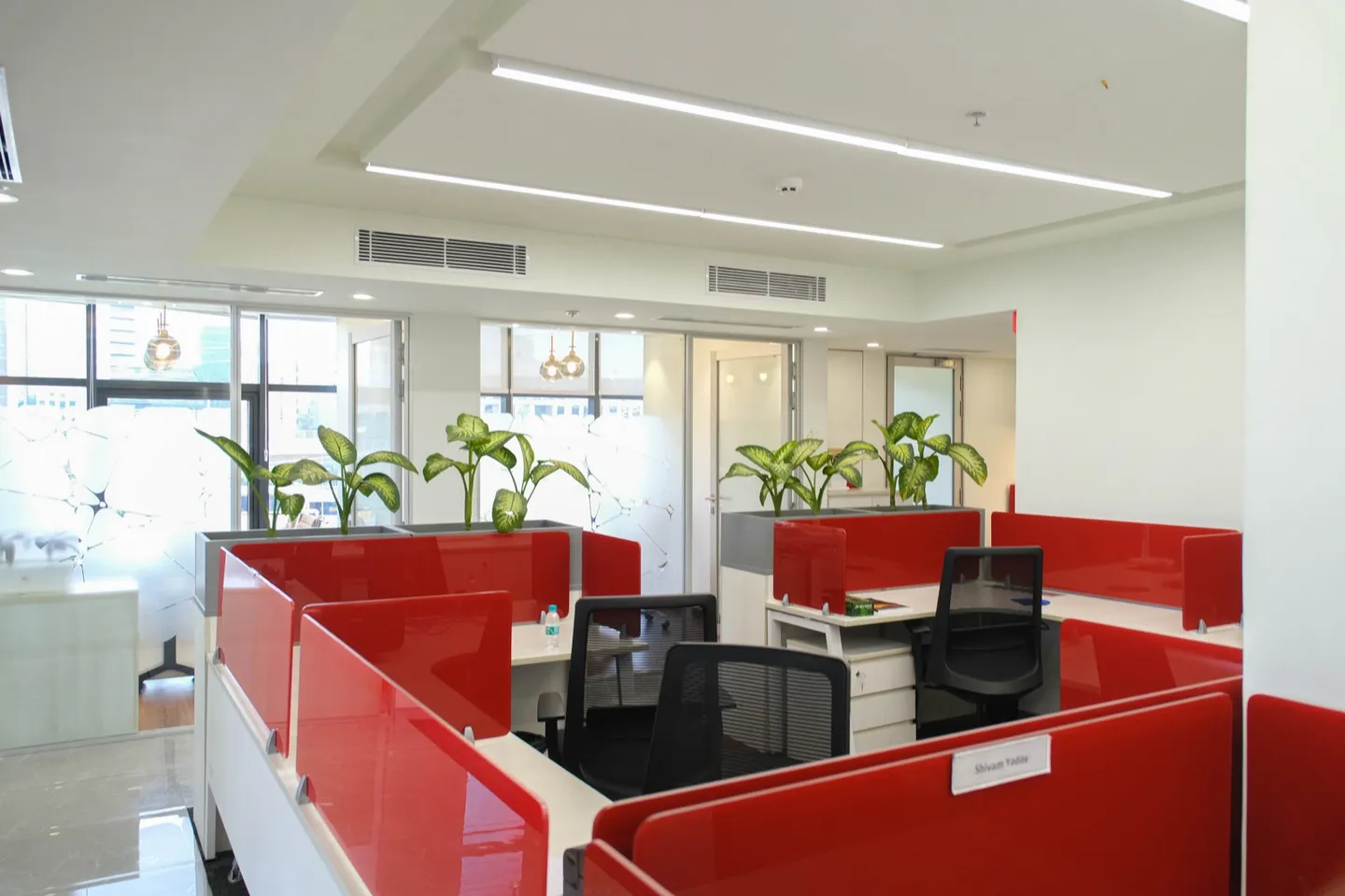 Desk cluster with red glass screens, planters and windows with city view — AIHP managed office, Gurgaon