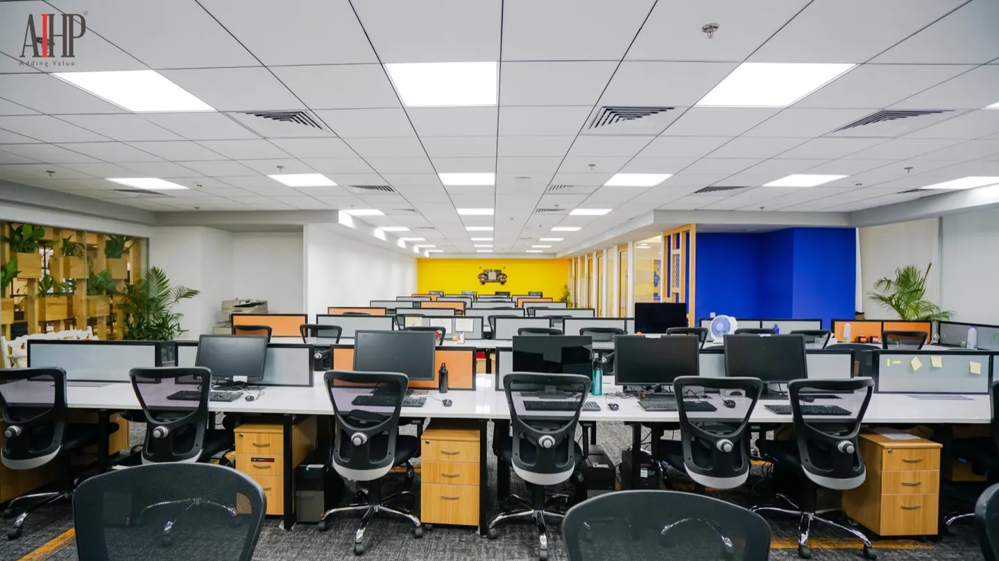 Desk rows with monitors and chairs by a yellow and blue accent wall — AIHP managed office, Gurgaon