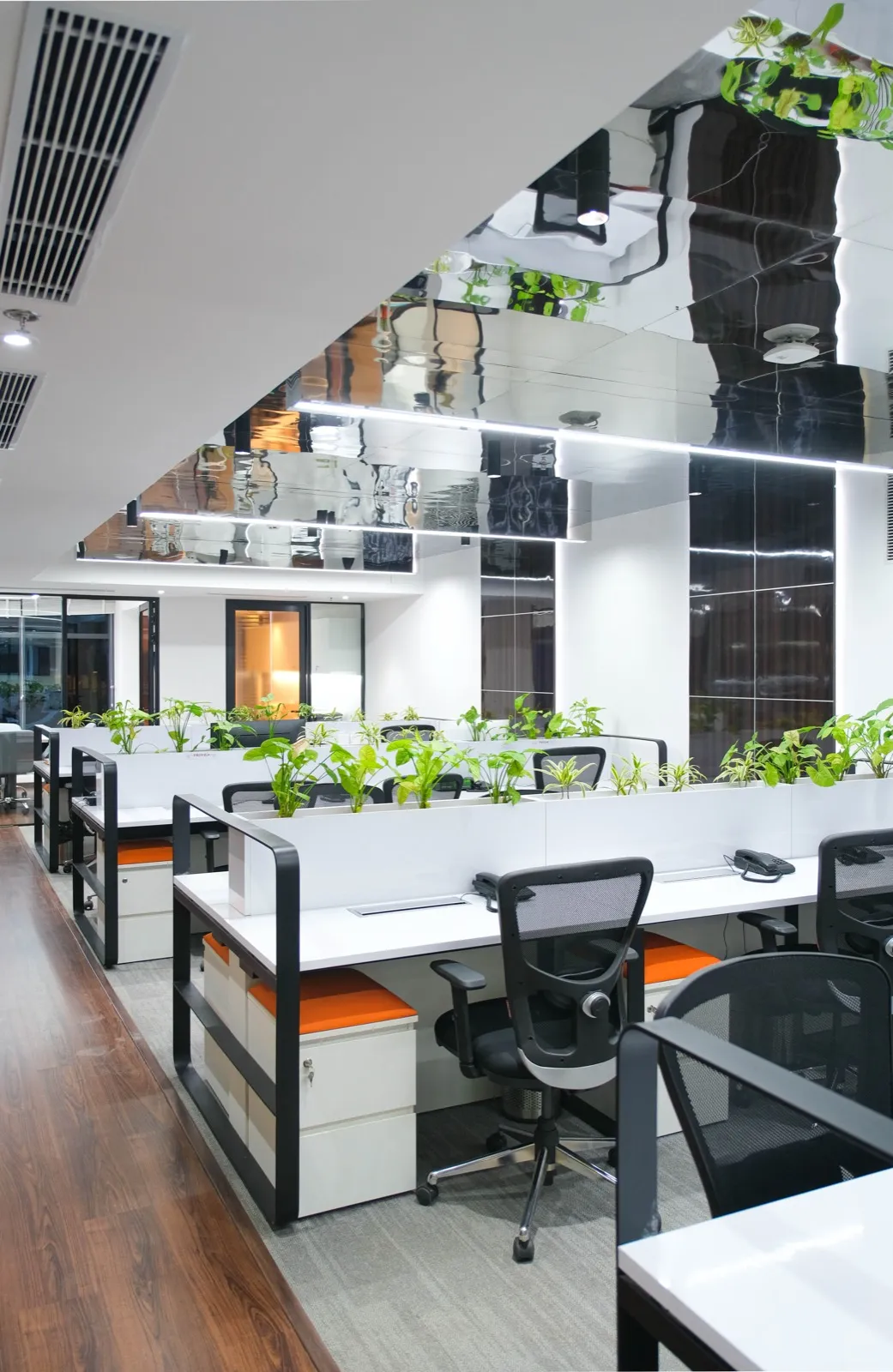 White desks with planters and orange seats below a glossy black ceiling band — AIHP managed office, Gurgaon