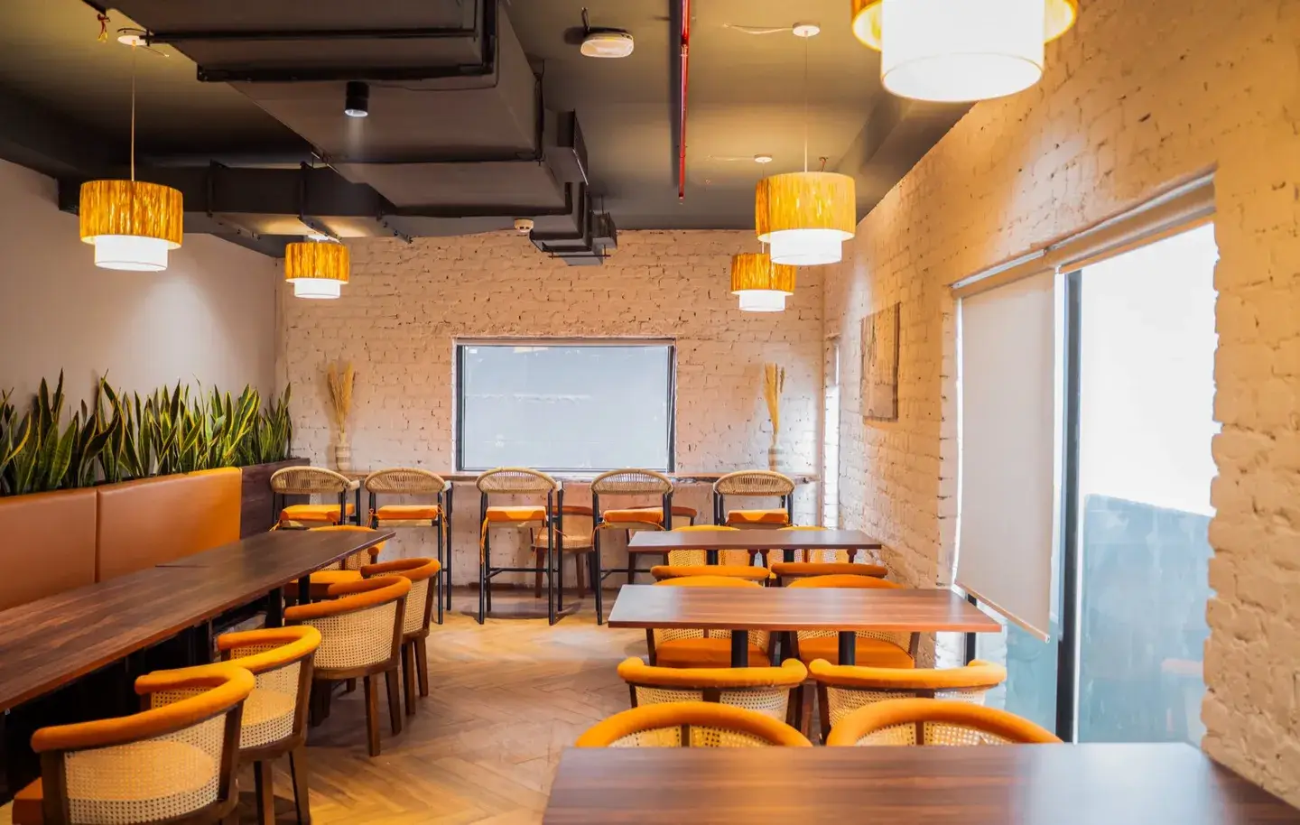 Breakout cafe with walnut tables, orange cane chairs and woven pendant lights on a whitewashed brick wall — AIHP managed office, Gurgaon